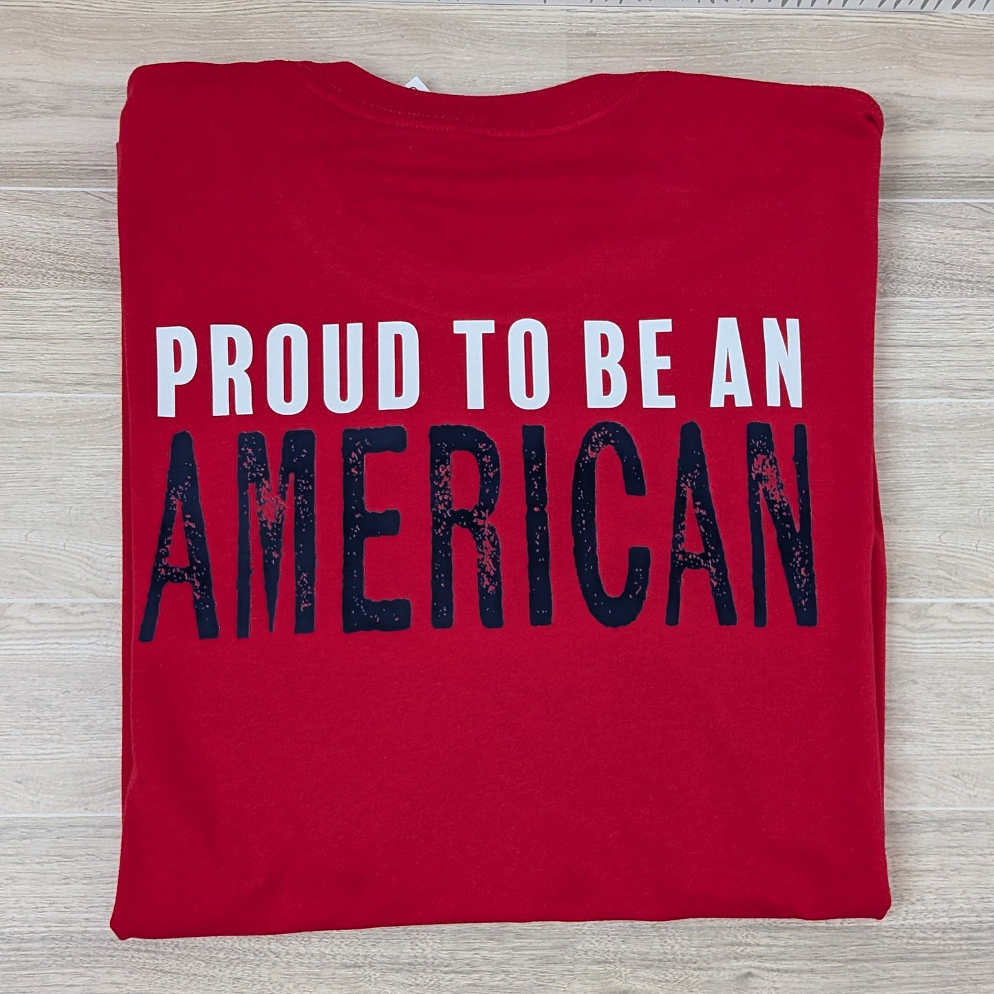 Red t-shirt with 'PROUD TO BE AN AMERICAN' text on a wooden surface